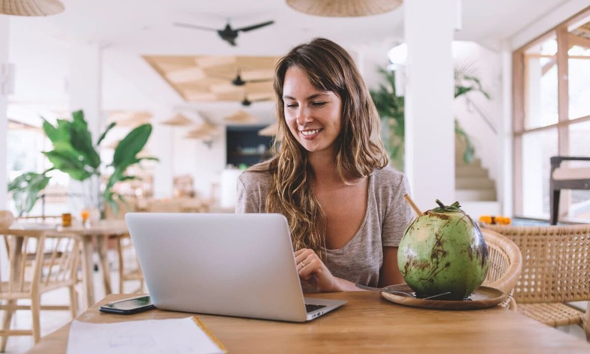 A digital nomad, working remotely on her laptop from a cafe in a tropical location, drinking from a coconut.